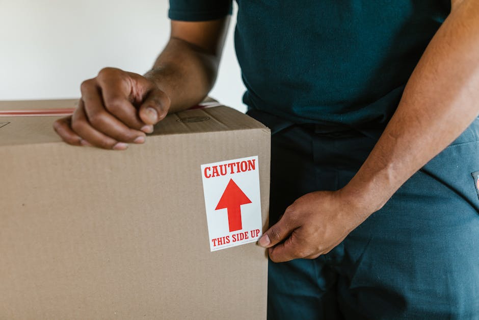 Close-up of a mover in dark clothing correctly handling a large cardboard box during a house relocation process. The mover is gripping the box with both hands, positioned at the sides, with a clear focus on maintaining stability and control during furniture transport. The box is sealed and features a prominent red and white caution label indicating 'CAUTION THIS SIDE UP,' which highlights proper handling procedures in packing and moving procedures. The background shows part of an indoor setting with a plain wall, and the scene captures the loading process inside a home, illustrating careful packaging and furniture transport by Movers Maida Vale as part of their removal services aimed at avoiding delays during house moves.