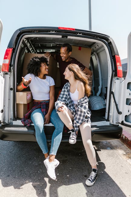 Three young adults sit inside the open cargo area of a large moving van, which is parked outdoors in daylight. They are engaged in conversation, smiling and appearing relaxed. The van's interior contains cardboard boxes and packing materials, indicating a home relocation process. The women are casually dressed, one wearing a white top and ripped jeans, the other in a checkered shirt and white dress, while the man stands behind them, leaning into the van. The scene captures the loading process or the final stages of packing for a house move, supported by the proximity to the van's rear door and the visible packaging materials. This setting reflects the logistics of furniture transport and packing in the context of professional removals, exemplified by companies like Movers Maida Vale, who assist with time-efficient moving services and relocation planning, aligning with the topic of booking delays and tips to avoid waits during house removals in Maida Vale.