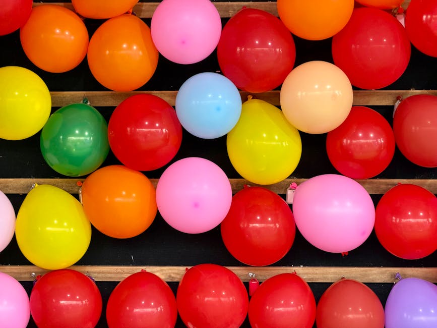 A display of multicoloured balloons in shades of red, orange, yellow, pink, green, blue, and purple, arranged on a black surface with wooden slats running horizontally across the image. The balloons are inflated and shiny, reflecting light, with some attached to strings or small weights at their bases. They are positioned in a somewhat random pattern, filling the frame from the top to the bottom, with no visible background or indoor environment. This colorful balloon arrangement may be part of a celebration or event setup, and the image showcases the variety of colours and the glossy texture of the balloons. The scene highlights the vibrant, festive atmosphere that could be associated with housewarming or moving events, relevant to professional removals as provided by Movers Maida Vale.