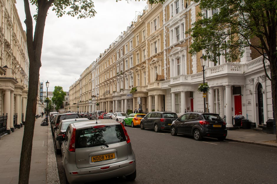 A residential street with a row of white Victorian-style terraced houses featuring detailed architectural elements, bay windows, and decorative facades. Several vehicles, including a grey van positioned on the left side and other cars parked along the curb, are visible, with the back of the van facing the viewer. Moving blankets and plastic wrapping are seen nearby, indicating ongoing home relocation or furniture transport. A person can be seen carrying a cardboard box through the open doorway of one of the houses, suggesting packing or unpacking activities. The street is lined with black iron railings, lampposts, and trees providing greenery amidst the rows of historic buildings under an overcast sky. This scene captures the typical setting for furniture loading or unloading during a house removal process, with visible effort and logistical arrangements associated with professional removals services like those offered by Movers Maida Vale.