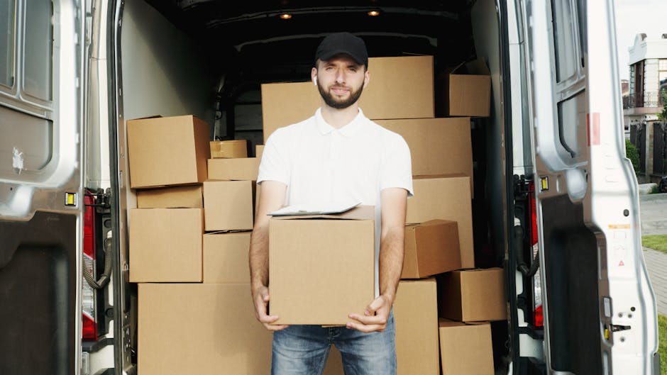 A young man wearing a white polo shirt and a black cap stands inside the open rear of a moving van, holding a cardboard box. The van's interior is filled with neatly stacked cardboard boxes of various
