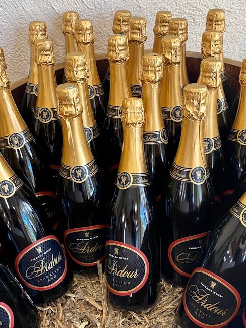 A close-up view of multiple dark glass bottles of Adduar sparkling wine with gold foil wrapping around the necks, arranged upright on a bed of crumpled packing paper inside a cardboard box. The bottles are part of a home relocation or packing process, evident from their arrangement and packing materials, which are visible behind a clean, neutral wall. The scene is lit evenly, highlighting the shiny foil and labels, and suggests careful handling typical of furniture and household items during moving services offered by Movers Maida Vale, particularly in a narrow streets setting such as Elgin Avenue W9.