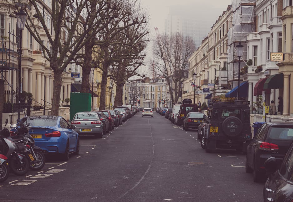 A narrow urban street scene in Maida Vale with parked cars along the right side and a white moving van in the middle, used for furniture transport during a home relocation. The pavement is lined with