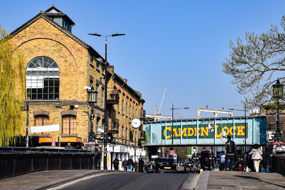 The image depicts a busy street scene in Maida Vale with a prominent brick building featuring large arched and rectangular windows, and a small rooftop structure. In the foreground, there are several pedestrians walking along the pavement near parked cars and a lamppost with mounted streetlights. A blue railway bridge crosses above the street, displaying the words 'Calden Lock' painted in large yellow letters on a weathered metal surface, indicating the nearby canal or railway line. Adjacent to the brick building, a shop sign for 'Orange Juice' is visible, with people standing outside. The street is lined with trees and urban infrastructure, under a clear blue sky, suggesting good daytime visibility. This scene may illustrate elements involved in house removals and furniture transport, such as loading zones, and the urban environment where professional movers like Movers Maida Vale operate for timely and efficient same-day relocations, avoiding hidden last-minute fees.