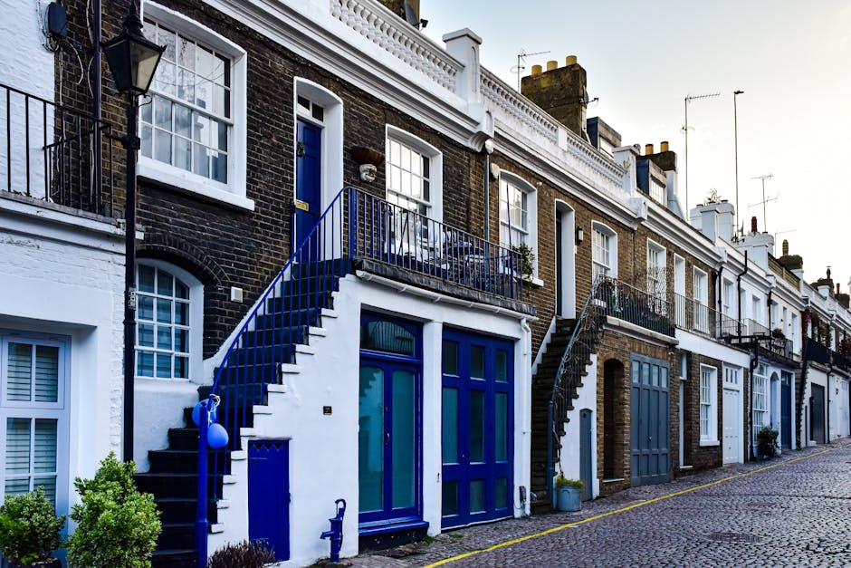 A row of traditional terraced houses with brick facades and white window frames, situated along a cobbled street in Maida Vale. Each property has a small front door and a staircase leading to the first-floor balcony, with some stairs featuring black or blue railing. The ground-level entrances include large glass doors, some with blue frames, and a few have potted plants or small shrubs nearby. The street is quiet and lined with parked vehicles, with a clear sky overhead, suggesting daylight. The scene captures the typical appearance of London residential streets, highlighting the staircase and parking tips relevant to house removals and furniture transport using professional services such as Movers Maida Vale.