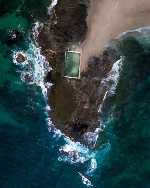 Aerial view of a coastal area showing a narrow strip of rocky shoreline with a concrete swimming pool situated on a small, elevated landmass connected to a sandy beach by a narrow pathway. The shoreline is surrounded by turquoise and dark blue ocean waters with visible waves crashing against the rocks and beach. The sandy beach extends along the right side of the image, with some vegetation visible at the top edge. The scene captures the natural landscape and coastal features adjacent to the water, with no visible human activity or vehicles present. This image illustrates a secluded seaside location, ideal for house removals or relocation services that include coastal property transfers or waterfront homes, as seen from the company Movers Maida Vale.