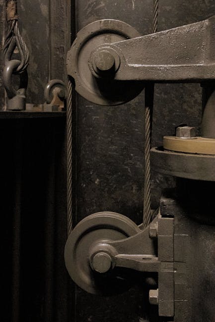 Close-up view of an industrial pulley system consisting of two large metal wheels connected by a thick steel cable, mounted vertically on a dark, textured metal wall. The system is part of a mechanical or lifting device used in moving or lifting heavy objects. In the background, there are other metal components and support structures, with minimal lighting highlighting the metallic surfaces. This image captures the detailed machinery involved in the loading process for house removals, as handled by Movers Maida Vale, illustrating the equipment used for safe furniture transport and furniture transport operations in a home relocation setting.