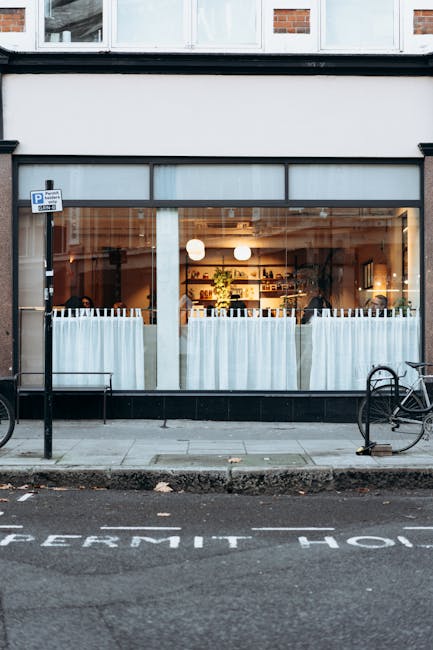 The image shows the exterior of a commercial property with large glass windows and a black frame, revealing the interior illuminated by warm lighting. Inside, there are shelves with various items, and a few potted plants are visible near the windows. Outside, on the pavement directly in front of the shop, there are two bicycles secured to metal bike racks, with one bike partially visible on the right side. The sidewalk is clean, with a curb separating it from the street, where a partially obscured marking reads 'PERMIT HO'. The scene is during daylight hours, with natural light softly illuminating the storefront. This setting could be relevant for property access or loadings during a house move, with Movers Maida Vale providing removals and furniture transport services, as indicated by the context of the webpage related to house removals and local moving regulations.